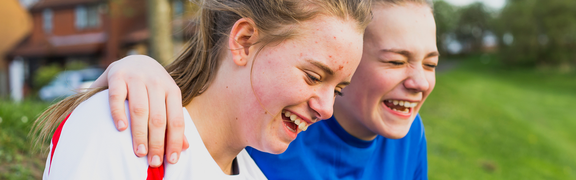 Close up of two teenagers laughing outside on a patch of grass. One wears a white t-shirt the other in a blue t-shirt. The latter has their arm on the other.
